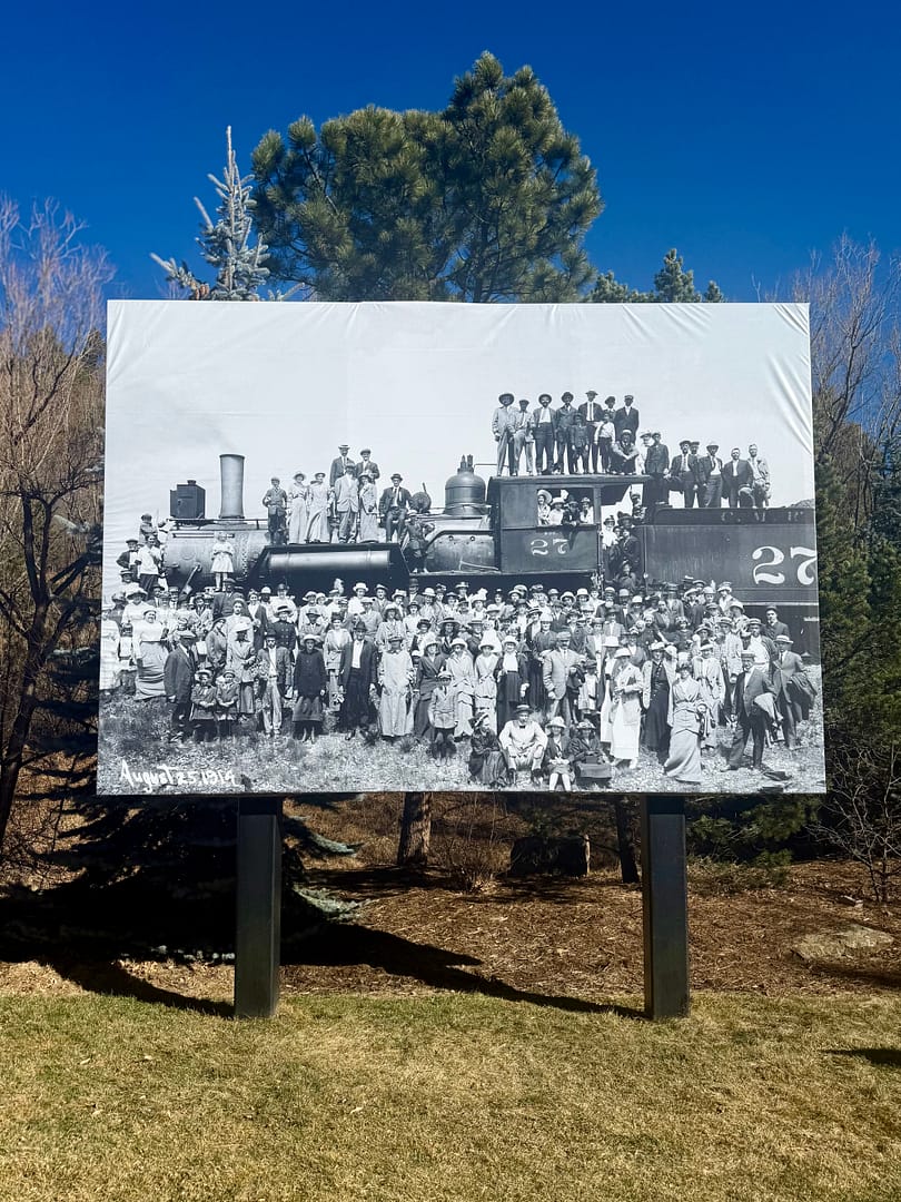 A black and white historical photo shows people gathered around an old train for a photograph.