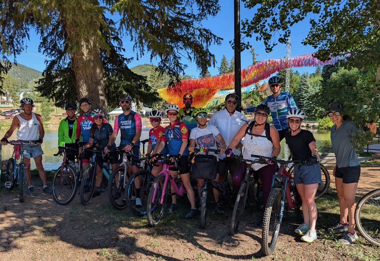 A group of people stand posing and smiling under a tree in the mountains