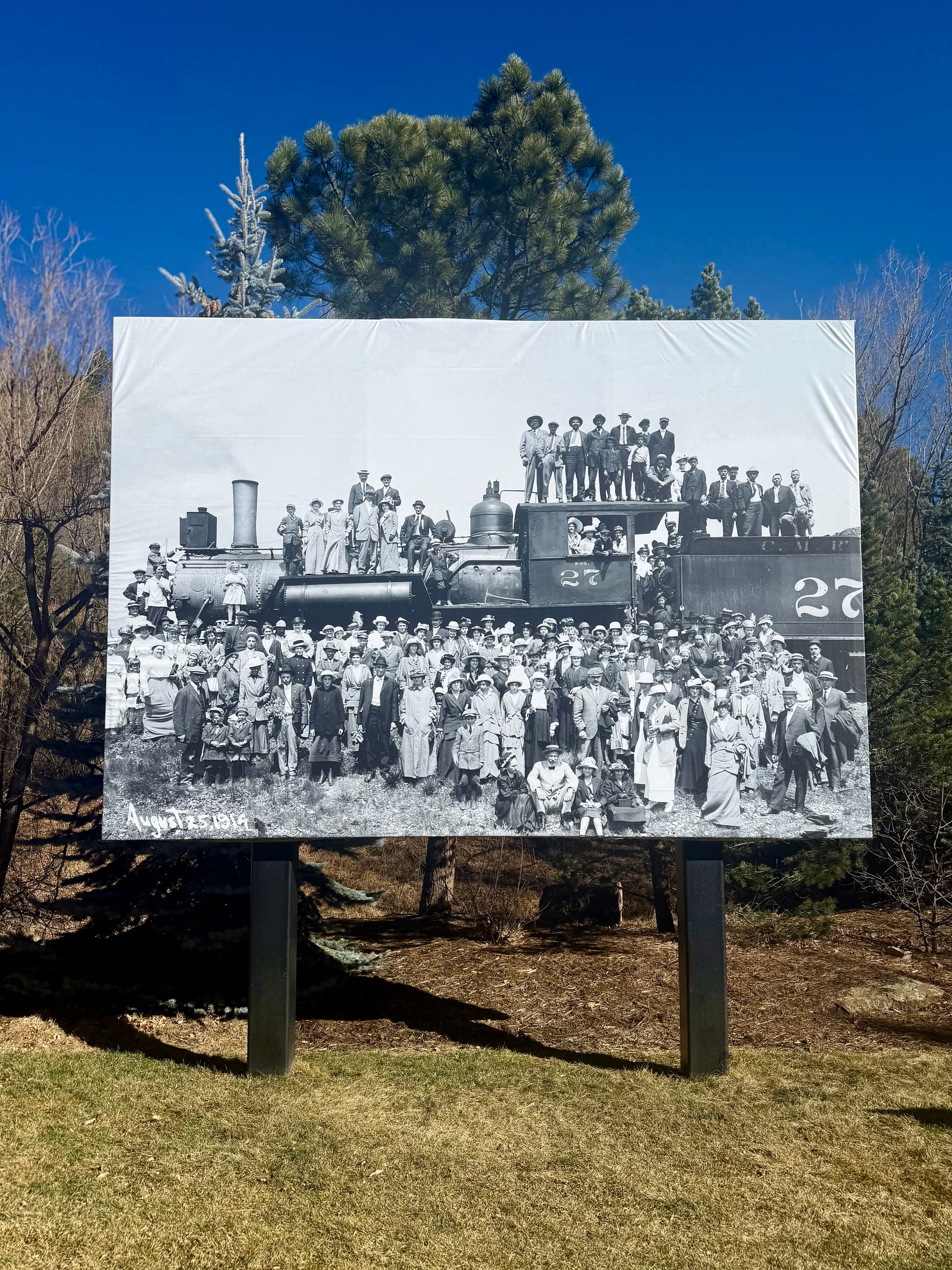 A black and white historical photo shows people gathered around an old train for a photograph.