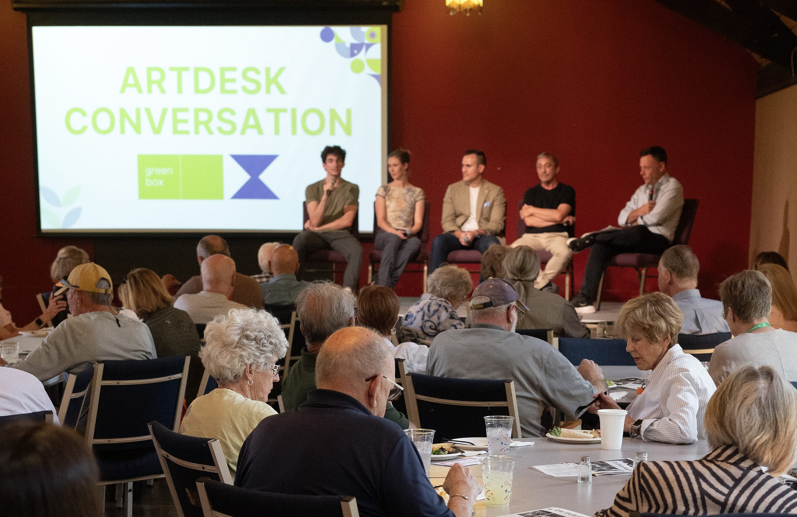 A group of people sit at tables eating lunch while listening to a panel of speakers sitting on a stage.