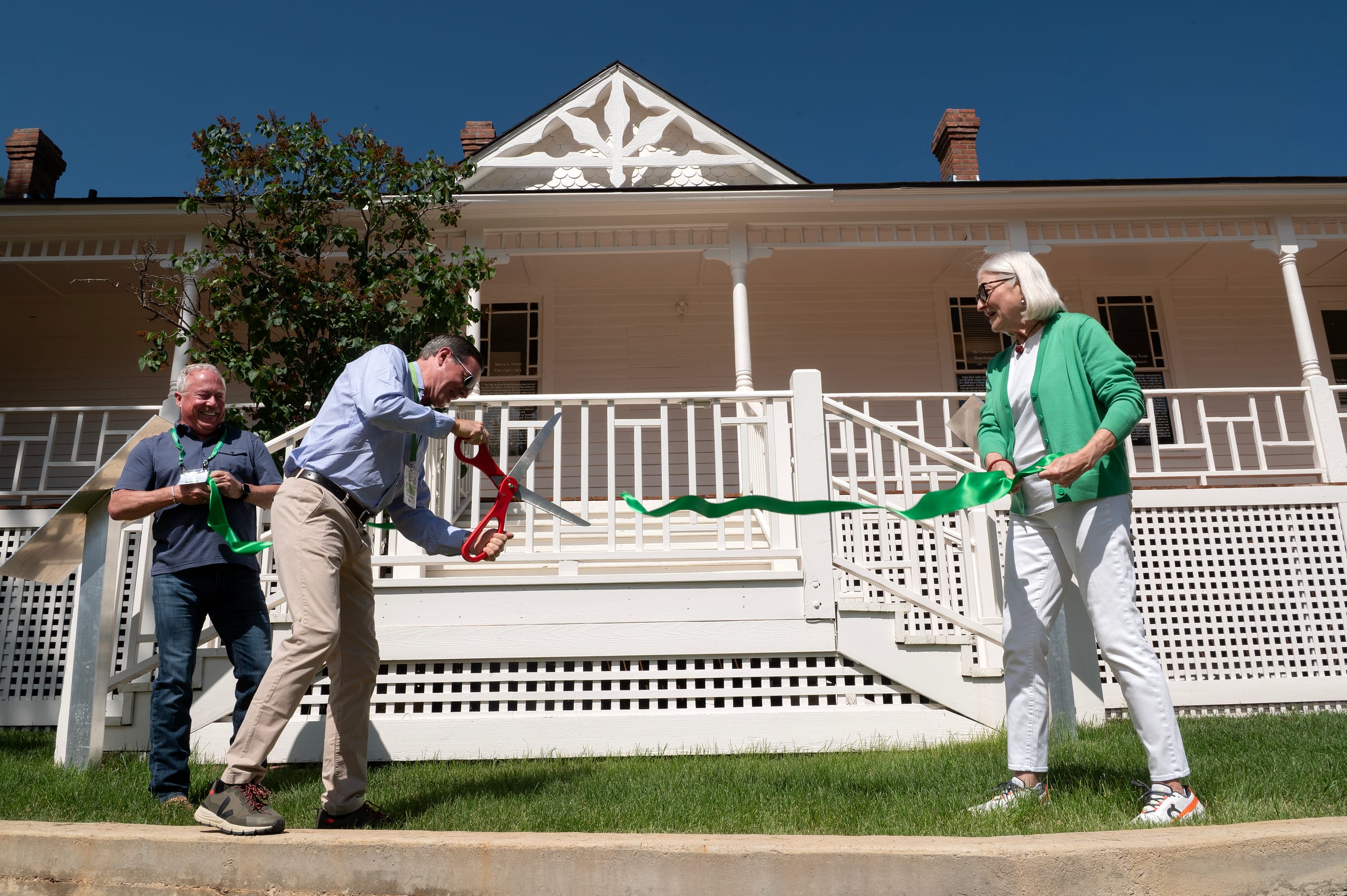 Three people stand in front of a white building. Two people hold a green ribbon while one person cuts the ribbon with large scissors.