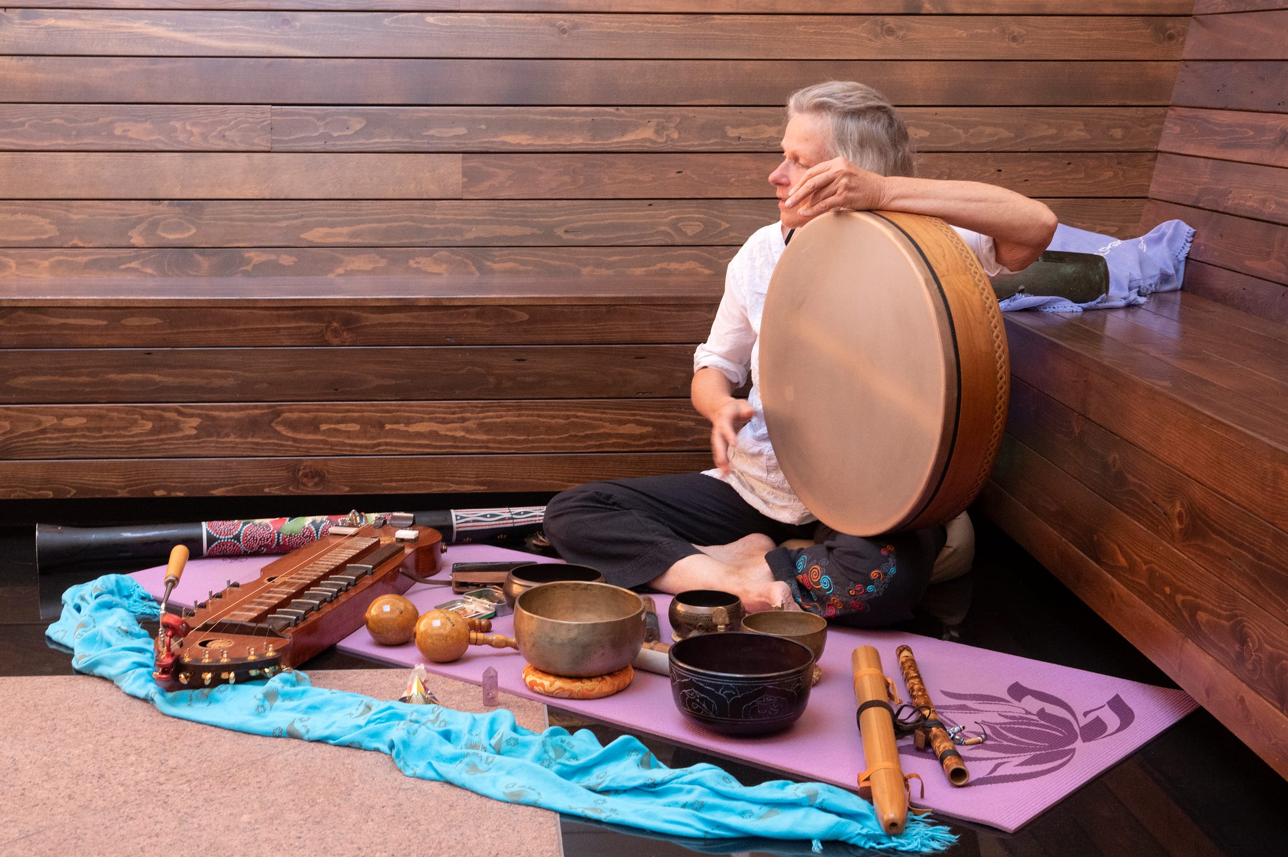 A woman sits inside a space with wooden walls. She sits on top of a blanket and has a range of instruments around her.