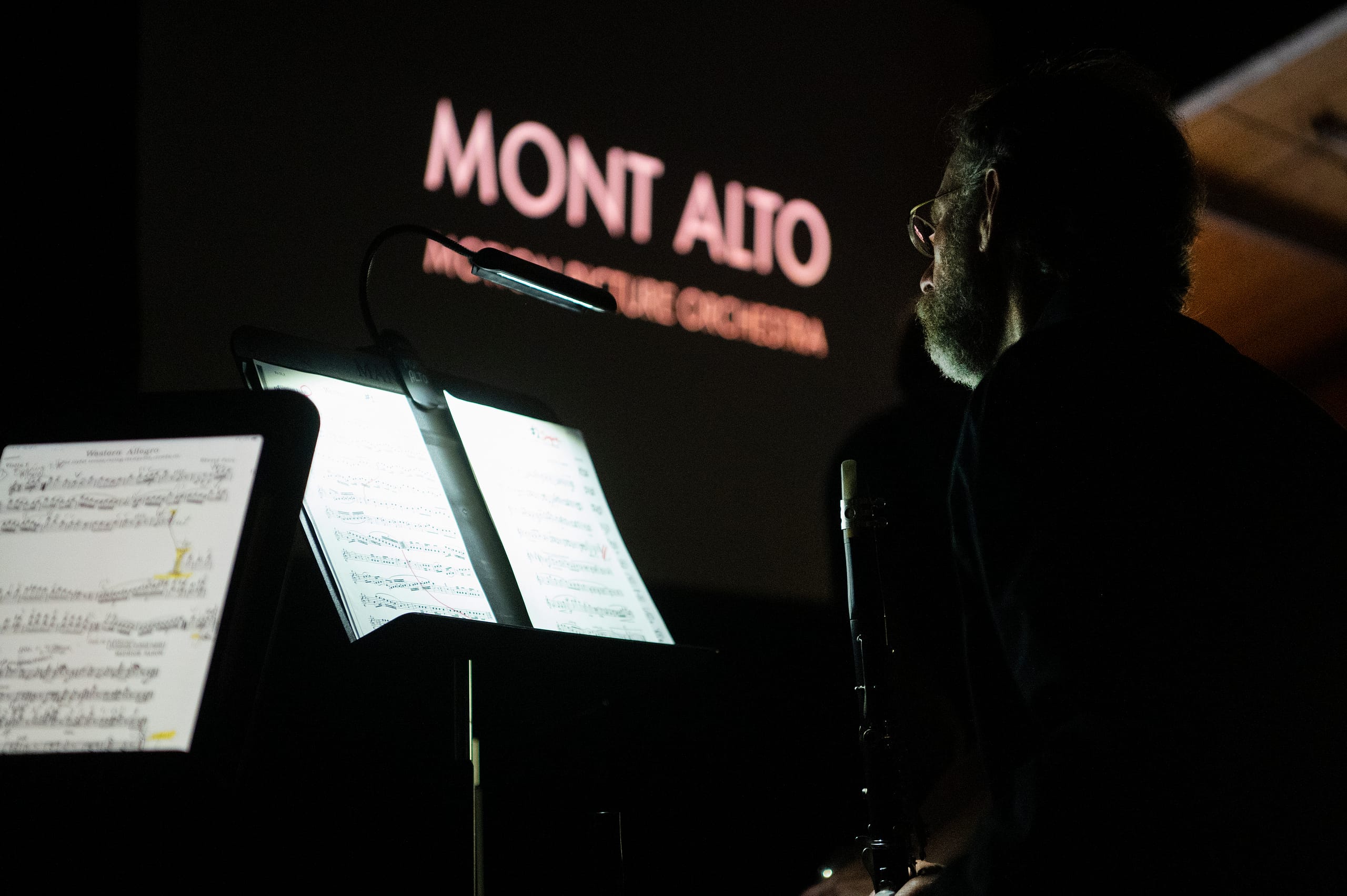 A man sits in front of a music stand that is illuminated. A sign in the background reads "Mont Alto."
