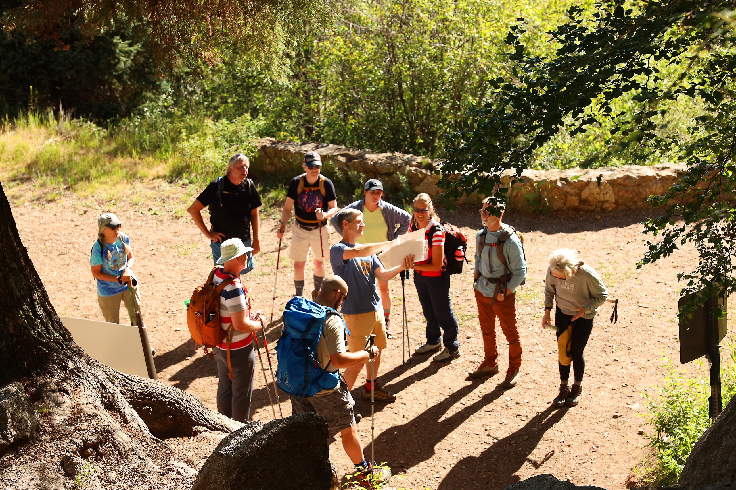 A man points out information on a map to a group of people that are standing around him. They are on a hiking trail on a sunny day.