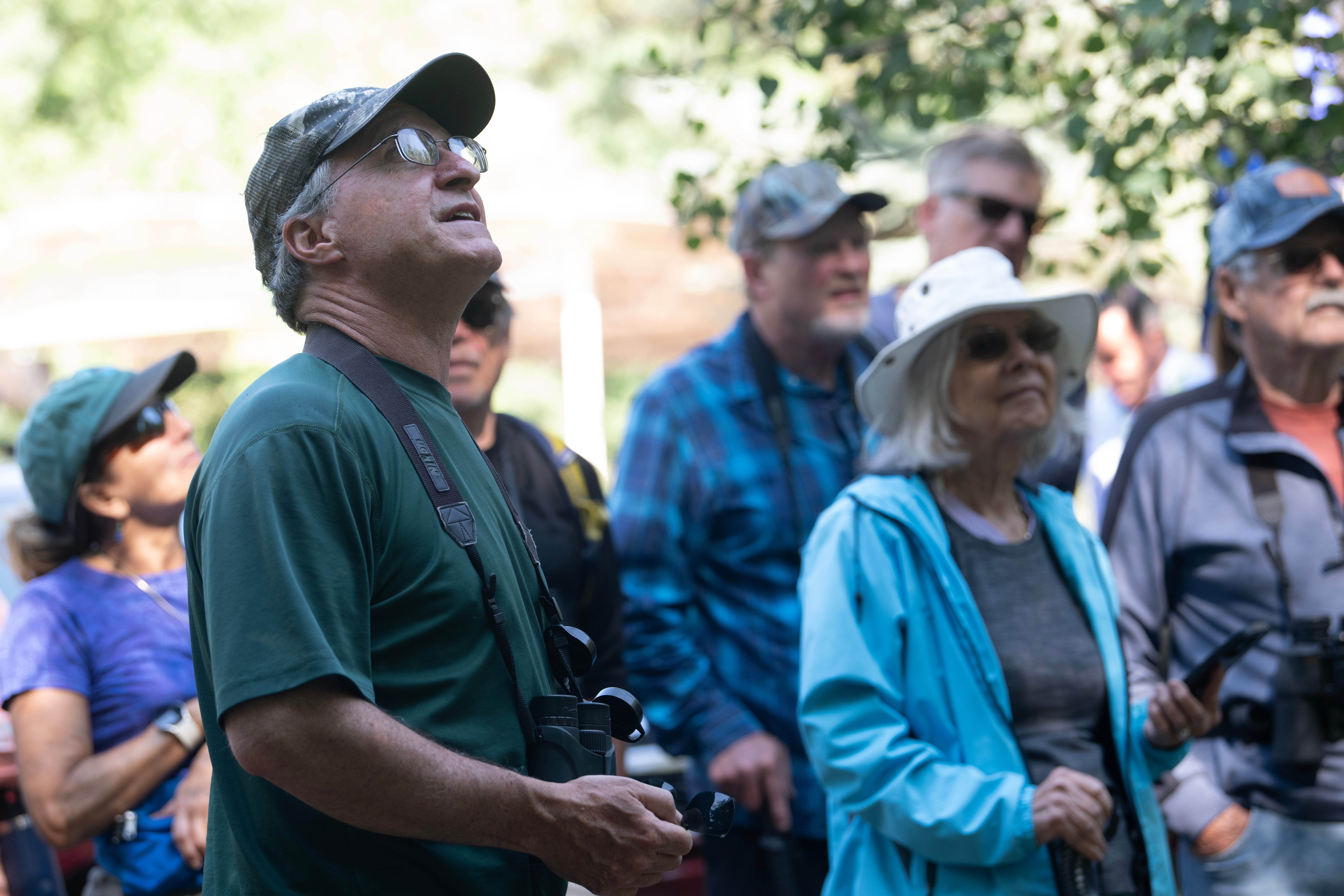 A man with binoculars stands next to a group of people looking up towards the trees